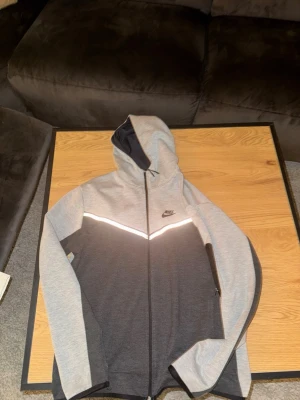 Nike hoodie i grått och svart - Nike hoodie i tvåfärgad design med ljusgrå upptill och mörkgrå nedtill. Reflekterande detalj över bröstet, hel dragkedja och huva med mörkblå insida. Perfekt för en sportig och avslappnad stil.