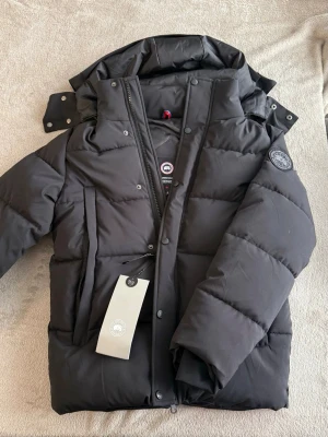 Svart dunjacka  - Säljer en svart dunjacka med avtagbar huva och klassisk logotyp på ärmen. Jackan har en rak passform, kraftig dragkedja och tryckknappar framtill. Perfekt för kalla vinterdagar och riktigt varm tack vare dunfyllningen.