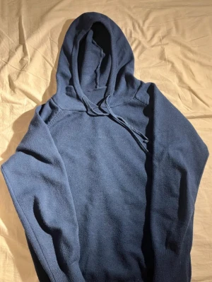 Mörkblå Kasmirhoodie  - Den är ganska liten i storleken. En enkel och stilren mörkblå hoodie med huva och dragsnören. Hoodien har ribbade muddar vid ärmslut och nederkant. Materialet ser ut att vara mjukt och lite tjockare, perfekt för att hålla värmen. Inga synliga loggor eller märken.