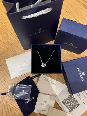 Swarovski halsband med svanberlock - Elegant halsband från Swarovski med en silverfärgad kedja och en gnistrande svanberlock dekorerad med vita och blå kristaller. Kommer i originalförpackning med certifikat och presentpåse. Perfekt för dig som gillar stilrena och exklusiva smycken.