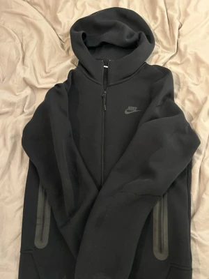 Svart Nike Tech Fleece hoodie - Nike Tech Fleece hoodie i svart med hel dragkedja och huva. Diskret Nike-logga på bröstet och två dragkedjeförsedda fickor på sidorna. Tillverkad i mjukt och lätt material som ger en clean och sportig look.