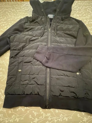 Svart pufferjacka från Polo Ralph Lauren - Snygg svart pufferjacka från Polo Ralph Lauren med quiltad front, huva med snörning och dragkedja hela vägen. Jackan har två fickor med tryckknappar och ribbade muddar vid ärmsluten. Perfekt för dig som vill ha en stilren och bekväm jacka.