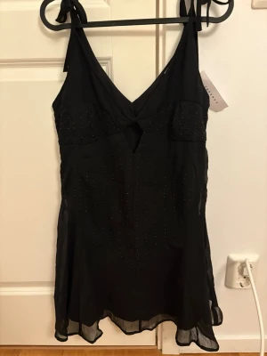 Svart klänning med strass och knytband - Snygg svart klänning med tunna axelband som knyts på axlarna. Klänningen har v-ringning, liten cut-out framtill och är dekorerad med strassdetaljer i vertikala rader. Materialet känns lätt och luftigt, perfekt för sommarkvällar. Klänningen är liten i storlek så skulle passa som en 38 också!!