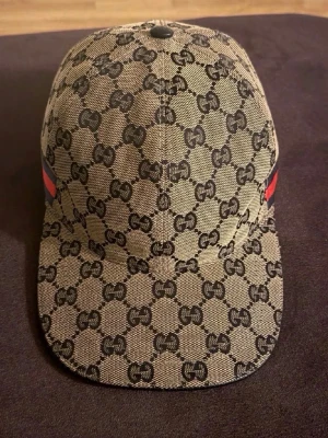 Navey Blue Gucci keps, med mönster - Säljer en beige keps från Gucci med klassiskt GG-monogram över hela kepsen och en röd-grön rand på sidan. Kepsen har böjd skärm och är tillverkad i canvasmaterial med svart knapp på toppen. Perfekt för dig som vill ha en ikonisk accessoar med lyxig känsla.