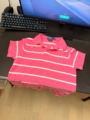 Rosa randig piké från Ralph Lauren ÄKTA!!!! - Säljer en rosa pikétröja från Polo Ralph Lauren med vita horisontella ränder och klassisk krage. Tröjan har korta ärmar och den ikoniska blå loggan broderad på bröstet. Perfekt för en avslappnad men ändå stilren look. Pris kan diskuteras!