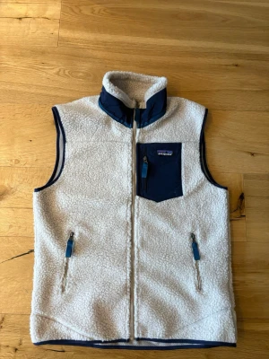 Vit fleeceväst från Patagonia - Snygg vit fleeceväst från Patagonia med marinblå detaljer. Västen har hög krage, hel dragkedja, två fickor med dragkedja och en bröstficka med logga. Materialet är mjuk teddyfleece och detaljerna är i slitstark nylon. Perfekt för lager-på-lager-stil.