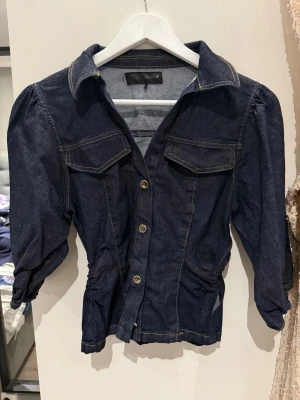 Mörkblå jeanstopp med puffärm - Snygg mörkblå jeansjacka med puffärmar och markerad midja. Jackan har två bröstfickor med lock, klassisk krage och knäppning framtill med metallknappar. Perfekt för dig som vill ha en trendig och unik jeansjacka.