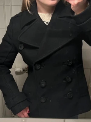 Svart skepparkavaj med dubbelknäppning - Stilren svart skepparkavaj med bred krage och klassisk dubbelknäppning framtill. Jackan har dekorativa axelklaffar och knappar vid ärmsluten. Materialet ser ut att vara ullblandning, vilket ger en snygg och tidlös look. Perfekt för dig som gillar klassiska plagg med modern touch.