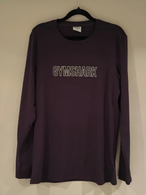 Svart långärmad topp från Gymshark - Långärmad svart topp från Gymshark med vit logotyp på bröstet. Perfekt för dig som gillar stilrena träningskläder och vill ha något bekvämt och snyggt. Materialet är mjukt och stretchigt, vilket gör den skön att bära både på gymmet och till vardags.