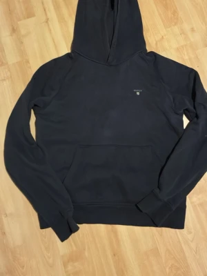 Svart hoodie från GANT - Svart hoodie från GANT med liten logga på bröstet. Klassisk modell med huva och känguruficka framtill. Tillverkad i mjukt material och har ribbade muddar vid ärmslut och nederkant. Perfekt för en avslappnad och stilren look.