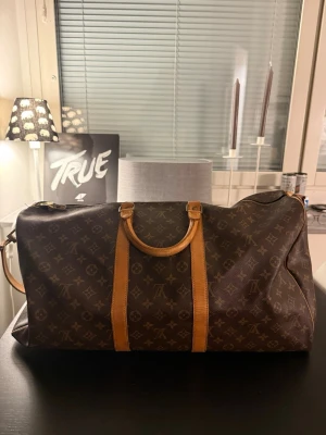 Louis Vuitton Keepall 55 Monogram väska - Säljer en klassisk Louis Vuitton Keepall 55 resväska i brunt monogrammönster med detaljer i ljust läder. Väskan har både handtag och justerbar axelrem, perfekt för weekendresor. Ikonisk design och rymlig insida för allt du behöver på resan.