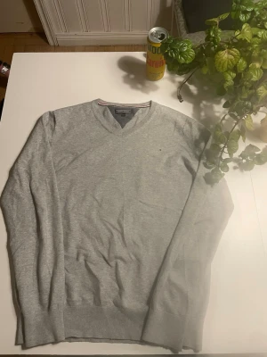 Grå V-ringad tröja Tommy Hilfiger - Snygg grå V-ringad tröja från Tommy Hilfiger med diskret logga broderad på bröstet. Tröjan har ribbade muddar vid ärmslut och nederkant. Perfekt för lager-på-lager och enkel att matcha med olika outfits.