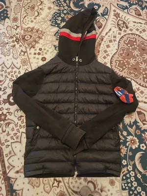 Svart pufferjacka med huva - Snygg svart pufferjacka med quiltad kropp och mjuka ärmar. Jackan har en huva med röda och grå ränder samt dragkedja framtill. Två sidofickor med knappstängning. Perfekt för kalla vinterdagar och riktigt chill stil, den är lite solbränd och dragkedjan går ner av sig själv när man går.