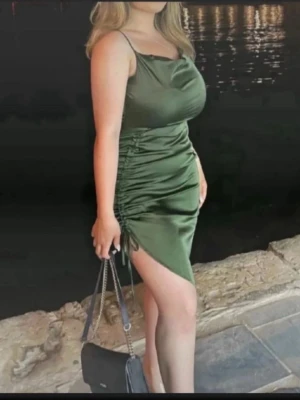 Grön satin klänning med snörning - Snygg grön kort klänning i satin med smala axelband och dragsko-snörning på sidan som ger en cool asymmetrisk look. Klänningen har en lätt glansig yta och figurnära passform, perfekt för dig som vill sticka ut.
