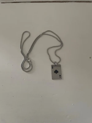 Halsband med ess i spader bricka - Snyggt halsband i silverfärgad metall med ett rektangulärt hänge som föreställer ett ess i spader från en kortlek. Kedjan är av klassisk modell och hänget har en blank finish med svart detalj. Perfekt för dig som gillar unika accessoarer med speltema.