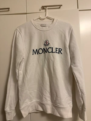 Vit Moncler sweatshirt med logga - Vit sweatshirt från Moncler med klassisk rund halsringning och stor Moncler-logga broderad på bröstet i svart. Tröjan har ribbade muddar vid ärmslut och nederkant, perfekt för en clean och stilren look.