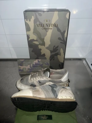 Grå kamouflage sneakers från Valentino - Snygga sneakers från Valentino i grått kamouflagemönster med vita och svarta detaljer. Skorna har vit/grå snören, mesh- och mockadetaljer samt en svart yttersula. Perfekta för dig som gillar grisch stilen och vill sticka ut i den stilen. BOX och allt finns med!!!!