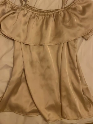 Champagnefärgad offshoulder topp i satin - Snygg offshoulder topp i champagnefärgat satinmaterial med volangdetalj upptill. Perfekt för dig som gillar en trendig och elegant look. Tunn och glansig finish som ger en lyxig känsla.