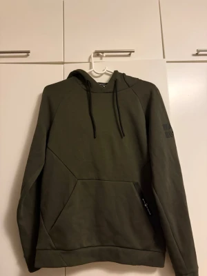 Olivgrön hoodie från Sail Racing - Snygg olivgrön hoodie från Sail Racing med svart dragsko i huvan och en stor magficka med dragkedja. Diskret logga på ärmen och coola sömdetaljer framtill. Perfekt för en avslappnad streetstil.