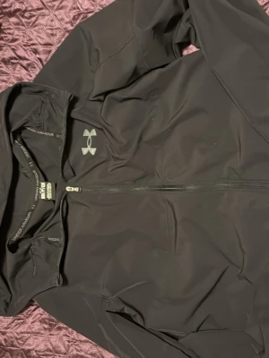 Svart vindjacka från Under Armour - Snygg svart vindjacka från Under Armour med dragkedja framtill och huva. Jackan har en stilren design med logga på bröstet och är tillverkad i lätt, vindtätt material som passar perfekt för aktiva dagar utomhus. Två jätte små hål i armen