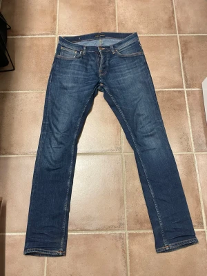 Blå jeans från Nudie  - Säljer ett par klassiska blå jeans från Nudie Jeans med raka ben och fem fickor och är modellen Grim Tim. Jeansen har snygga slitningar och kontrastsömmar samt Nudie-logga på bakfickan. Tillverkade i bomull med en tidlös tvätt som passar till det mesta.
