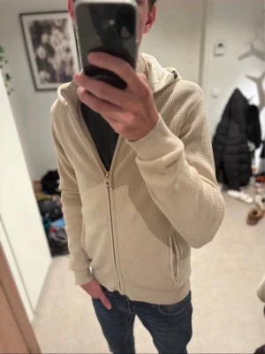 Indicode cardigan S - Säljer en beige hoodie med dragkedja framtill och två fickor med dragkedja. Hoodien har ribbade muddar vid ärmslut och nederkant samt en huva. Materialet är ribbstickat och ger en avslappnad look. Perfekt för dig som gillar stilrena och enkla plagg.