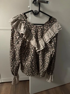 Blommig volangblus med långa ärmar - Supersöt blus med svart botten och rosa blommönster, dekorerad med en bred volang över axlarna i kontrasterande vitt småblommigt tyg. Långa ärmar med resår vid ärmslut ger en puffig effekt. Perfekt för dig som gillar romantisk och vintage-inspirerad stil.