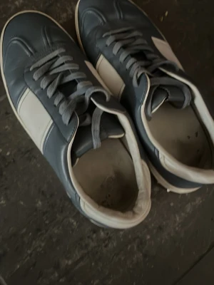 Gråvita sneakers med retrostil - Snygga sneakers i grått och vitt med klassisk retrodesign. Skorna har vita sidor, grå ovandel och matchande grå skosnören. Tillverkade i slätt konstläder med lågt skaft och vit sula. Perfekta för dig som gillar stilrena och tidlösa sneakers.