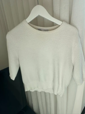 Vit stickad tröja från Zara - Säljer en enkel och stilren vit stickad tröja från Zara. Tröjan har rund halsringning, korta ärmar och ribbade kanter. Perfekt att matcha med jeans eller kjol för en clean look. Materialet känns mjukt och bekvämt.