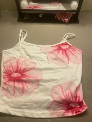 Vit linnetopp med rosa blommor - Säljer en söt vit linnetopp med tunna axelband och stora rosa blommor som tryck. Toppen har en enkel och fräsch design, perfekt för varma dagar. Materialet känns mjukt och lätt mot huden.