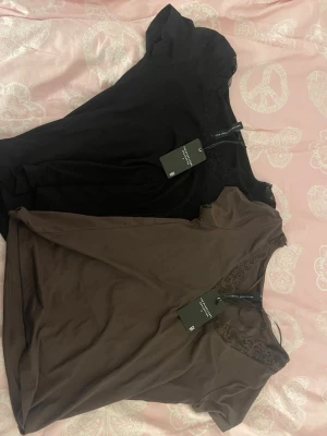 2 spetsdetaljerade toppar 157 Black Label - Två snygga toppar från 157 Black Label Collection, en svart och en brun. Båda har v-ringning med spetsdetaljer vid halsen och korta ärmar. Perfekta att styla till jeans eller kjol för en trendig look.