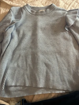 Ljusblå stickad tröja från Vila - Snygg ljusblå stickad tröja från Vila med ribbad struktur och volangdetaljer vid axlarna. Tröjan har rund halsringning och långa ärmar, perfekt för dig som vill ha en stilren men ändå trendig look. En fläck men går nog att tvätta bort.