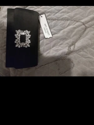 Svart clutch väska med kristaller London street art - Snygg svart kuvertväska från London Art Street med glittrande strassdetalj på framsidan och silverfärgad kedja. Väskan har magnetlås och är tillverkad i ett slätt material som ger en elegant look. Perfekt för dig som vill sticka ut lite extra.