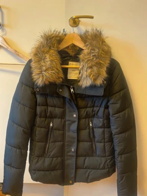 Svart pufferjacka med fuskpäls - Snygg svart pufferjacka med stor krage i beige fuskpäls. Jackan har två dragkedjeförsedda fickor framtill och quiltad design. Perfekt för kalla dagar och ger en trendig look. Dragkedja och knappar framtill för extra skydd mot kyla. Y2k