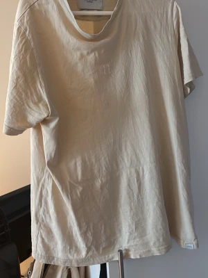 Beige t-shirt från Les Deux - Snygg beige t-shirt från Les Deux med diskret broderad logga på bröstet. Klassisk rund halsringning och avslappnad passform. Tillverkad i mjuk bomull som känns skön mot huden. Perfekt basplagg till garderoben. Har ett litet litet hål vid etiketten i tröjan vid kragen samt vad som upplevs som smuts/fläck på framsidan men syns inte på bild!