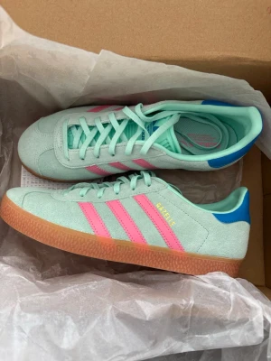 Adidas Gazelle mintgrön/rosa sneakers - Aldrig använda! Mintgröna Adidas Gazelle sneakers i mocka med klassiska rosa ränder på sidorna och blå detalj vid hälen. Skorna har gummisula i brun ton och snörning framtill. Perfekt till våren!
