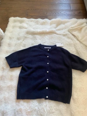 Mörkblå kofta med pärlknappar från lager 157 - Kortärmad mörkblå kofta från lager 157 med snygga pärlknappar framtill. Koftan har rund halsringning och är gjord i ett mjukt stickat material som känns skönt mot huden. Perfekt att slänga över en topp för en stilren look.