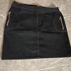 Svart manchesterkjol med dragkedjor - Snygg svart kjol i manchester med två framfickor som har dekorativa dragkedjor med ringdetaljer. Kjolen har en rak passform och är perfekt för att skapa en cool och trendig look.