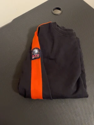 Svart hoodie med orangea ränder - Cool svart hoodie med tydliga orangea ränder längs ärmarna och ett P.I.S-märke på ena armen. Tröjan har ribbade muddar och är tillverkad i mjukt material, perfekt för en avslappnad streetwear-look.