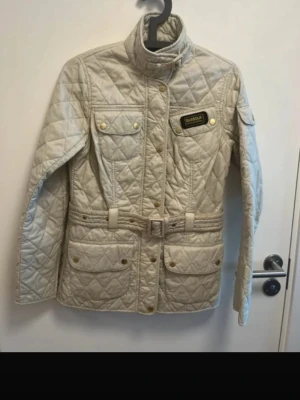 Beige quiltad jacka från Barbour - Stilren beige quiltad jacka från Barbour med bälte i midjan, hög krage och guldfärgade knappar. Jackan har fyra fickor framtill, Barbour-logga på bröstet och lång ärm. Perfekt för dig som vill ha en klassisk look.