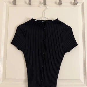 Svart ribbad topp från Gina Tricot - Säljer en svart ribbad topp från Gina Tricot med korta ärmar och knappar framtill. Toppen har en slim passform. Har storlek M i den men den är liten i storleken. Aldrig användt den.