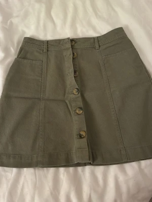 Olivgrön kjol med knappar framtill - Snygg olivgrön kjol i bomull med raka sömmar och fickor framtill.