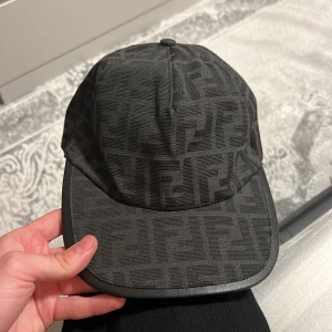 Svart keps från Fendi med monogram - Säljer en svart keps från Fendi med klassiskt FF-monogram över hela kepsen. Justerbar baktill med broderad Fendi-logga. Kepsen är tillverkad i ett tygmaterial och levereras med original dustbag.taggen har börjat suddas lite