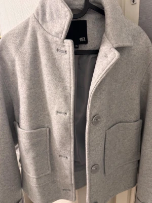 Ljusgrå kappa - Stilren ljusgrå rock från 157 i ullmix med rak passform och stora utanpåfickor. Rocken har hög krage, knäppning med stora knappar och slät insida. Perfekt för dig som gillar minimalistisk och modern stil. Använd 1 gång. 