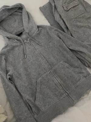 Grått Juicy Couture loungewear-set - Mysigt grått loungewear-set från Juicy Couture i  velour. Setet består av en zip-hoodie med fickor och dragsko samt matchande byxor med ficka bak. Klassisk Juicy-dragkedja och detaljer i silver. Tröjan är i s och byxorna i m