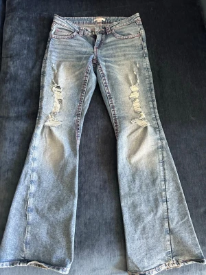 Jeans Gina tricot  - Säljer ett par ljusblå bootcut jeans med slitna detaljer på låren och knäna. Jeansen har fem fickor, röd kontrastsöm och klassisk knappgylf. Perfekta för dig som gillar en avslappnad och trendig look.