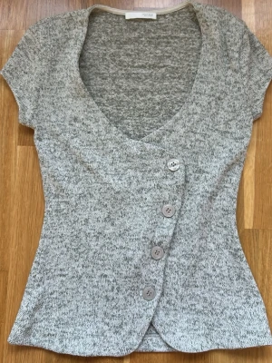 Gråmelerad topp med knappar från Zara - Kortärmad topp från Zara i gråmelerat stickat material. Djup V-ringning och fyra stora knappar framtill i en sned rad. Figurnära passform och mjuk, stretchig kvalitet. Perfekt till jeans eller kjol för en trendig look.