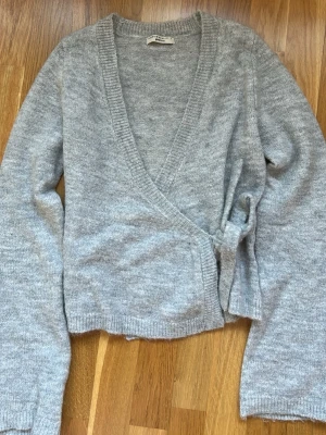 Grå omlottkofta från Gina Tricot - Säljer en mjuk grå stickad omlottkofta från Gina Tricot. Koftan har långa ärmar, ribbade kanter och knytdetalj i sidan. Perfekt att slänga över en topp för en chill look. Materialet är skönt och passar bra till lager-på-lager.