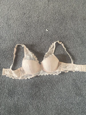 Beige spets-bh med vaddering - Säljer en beige bh med vadderade kupor och fina spetsdetaljer. Bh:n har justerbara axelband och hakknäppning bak. Spetsen ger en romantisk känsla och materialet är mjukt och bekvämt mot huden.