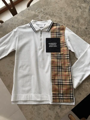 Vit Burberry henley med rutig detalj - Säljer en vit långärmad henley från Burberry med klassisk rutig panel i beige, svart och rött på ena sidan. Skjortan har svart patch med texten 'BURBERRY LONDON ENGLAND' på bröstet, krage med svarta ränder och half zip framtill. Perfekt för dig som gillar statement-plagg.